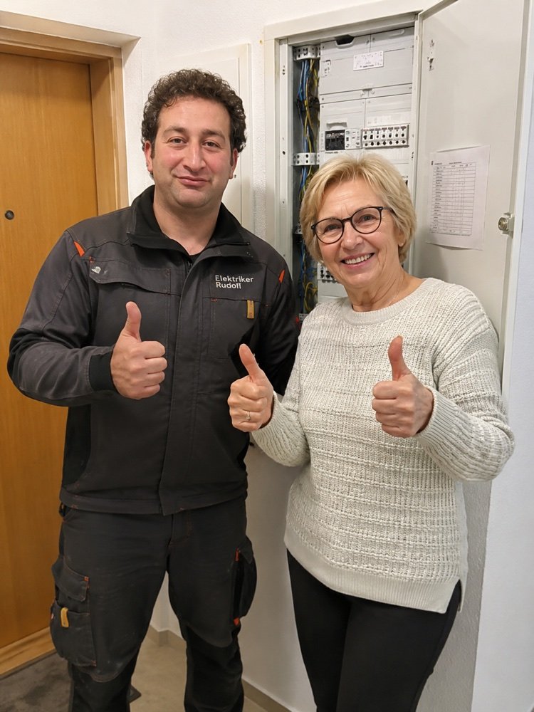 Zufriedene Kundin mit Elektriker Rudolf vor dem reparierten Sicherungskasten – beide zeigen Daumen hoch